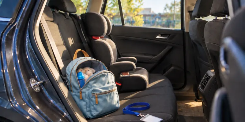 Through the open rear door of a family car on a bright autumn afternoon: a child's booster seat and a small blue backpack on the back seat, with a lanyard and a blank card tossed beside them.